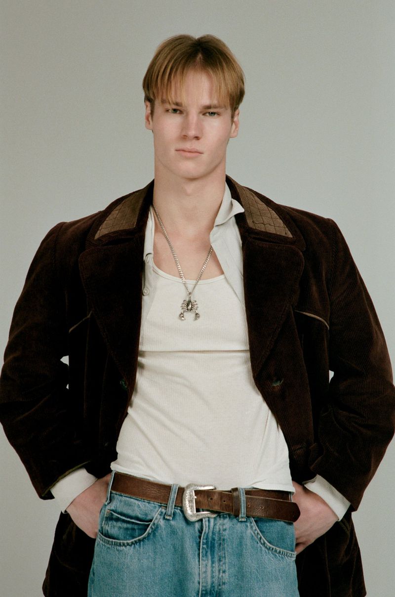 Model Henry Rank wearing a brown corduroy jacket, white tank top, and blue jeans against a grey background.