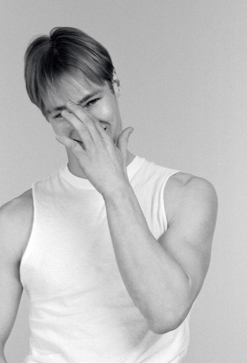 Black and white portrait of a smiling adult male with blonde hair wearing a white tank top against a solid light gray background.