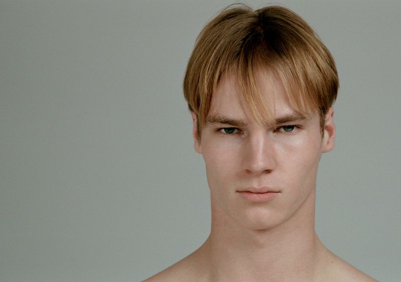 Close-up portrait of a young male model with short parted blond hair against a plain grey background