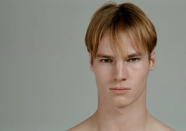 Close-up portrait of a young male model with short parted blond hair against a plain grey background
