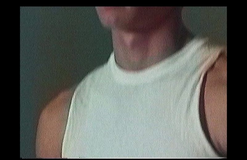Close up crop of a man wearing a white tank top against a dark green textured wall background.