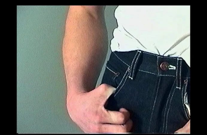 Close-up of model Henry Rank wearing dark jeans and a white top against a solid neutral wall background.