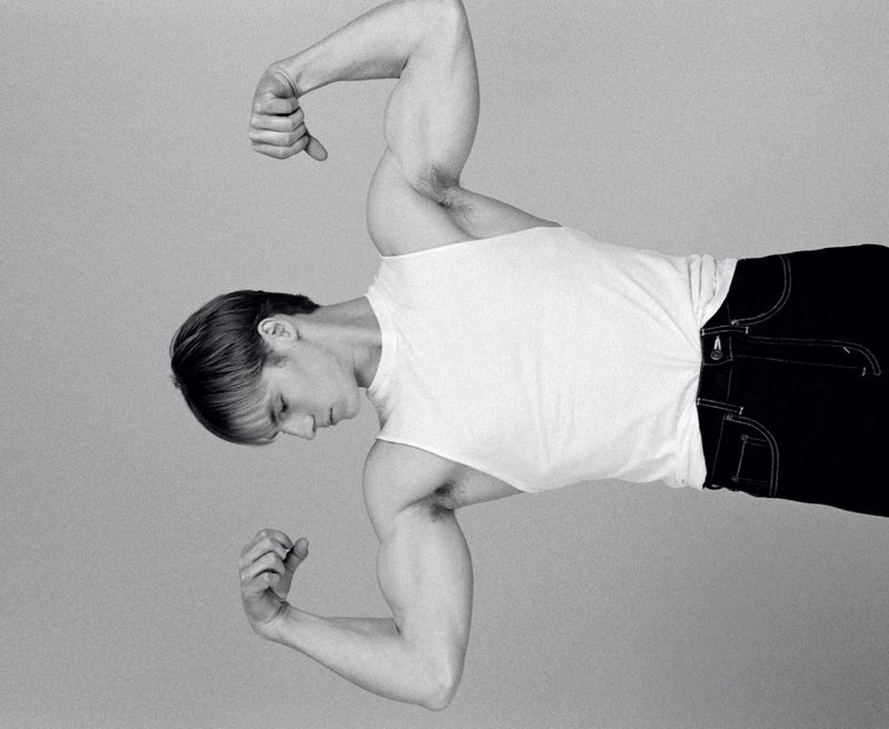 Model Henry Rank posing in a white tank top and dark pants against a seamless grey backdrop for HERO USA.
