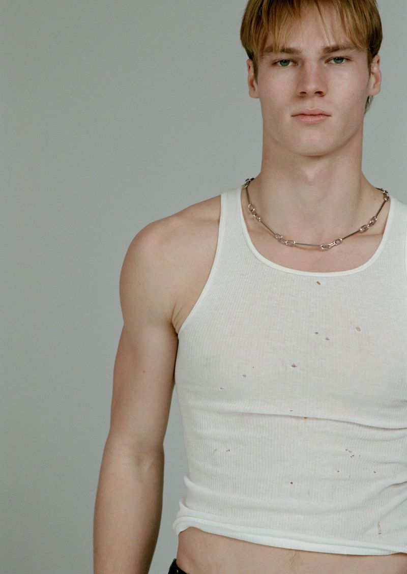 Portrait of male model Henry Rank wearing a white tank top and chain necklace against a neutral grey studio background
