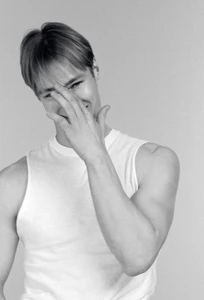 Portrait of male model Henry Rank in a white tank top, smiling with hand covering his face against a grey studio backdrop.
