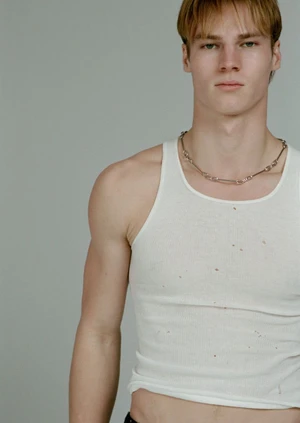 Portrait of male model Henry Rank wearing a white tank top and chain necklace against a neutral grey studio background