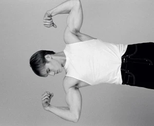 Model Henry Rank posing in a white tank top and dark pants against a seamless grey backdrop for HERO USA.