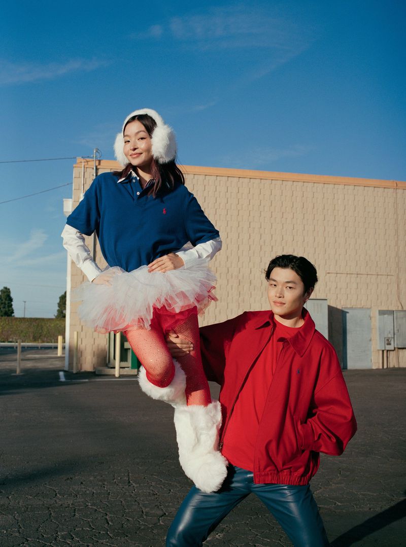 Man lifting a woman wearing a blue polo, white earmuffs, tutu, and furry white boots outdoors against a tan building.