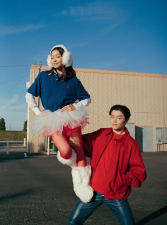 Man lifting a woman wearing a blue polo, white earmuffs, tutu, and furry white boots outdoors against a tan building.