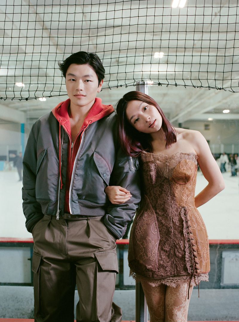 An Asian man and woman pose together indoors in an ice rink. The man wears a gray jacket with brown pants, and the woman wears a brown lace dress.