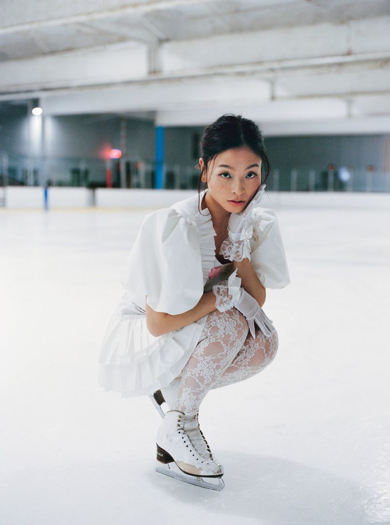 Asian woman in a white dress and ice skates crouching on an indoor ice rink