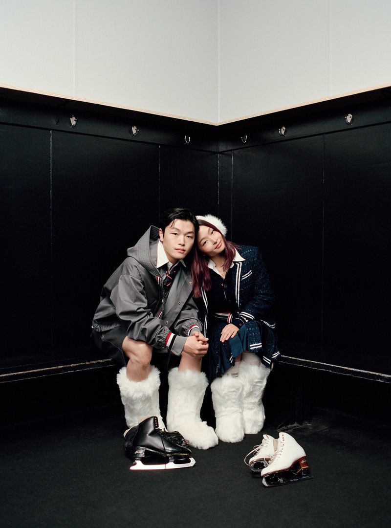 Two people wearing white fluffy boots seated against a dark locker room wall with ice skates on the floor
