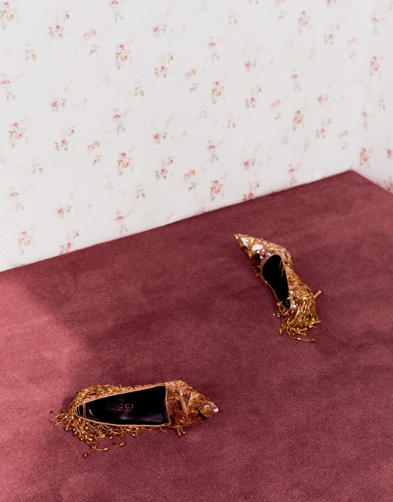 Two embellished gold stiletto heels resting on a plum carpet against a patterned wallpaper wall