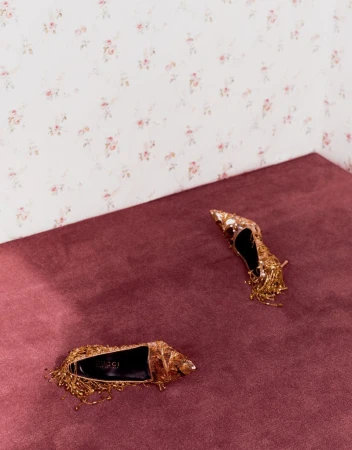 Two embellished gold stiletto heels resting on a plum carpet against a patterned wallpaper wall