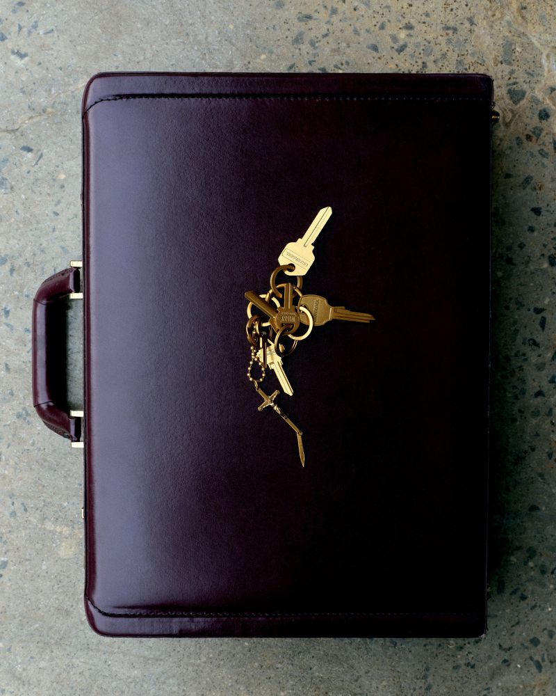 Top-down view of a dark burgundy leather briefcase resting on a textured grey concrete ground with a cluster of gold keys on top.