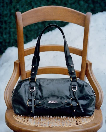 A black leather shoulder bag with straps and metal hardware resting on a wooden chair outdoors with snow in the background.