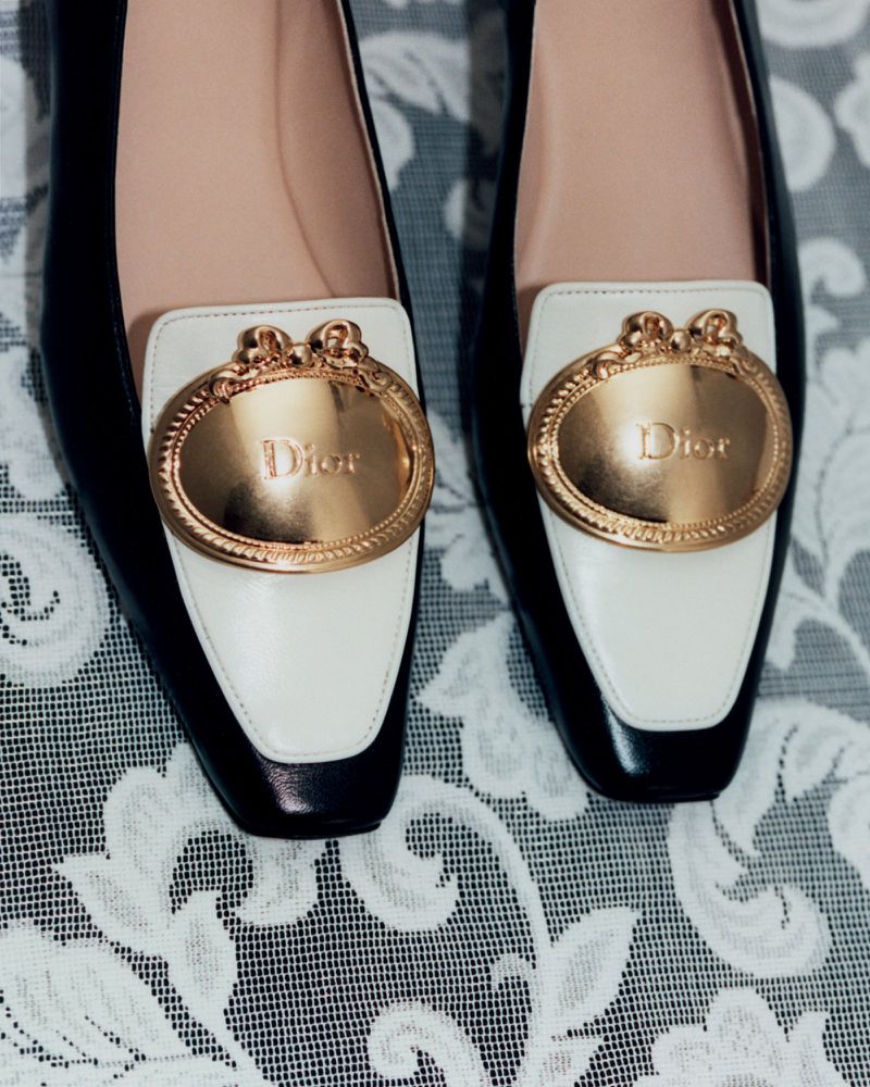 Close-up of black and white leather loafers featuring ornate gold Dior buckles resting on white lace fabric.