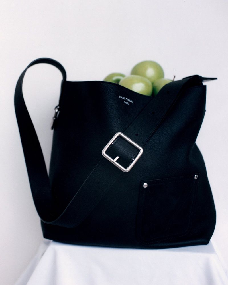 Black leather shoulder bag with a silver buckle and strap holding green apples against a white seamless background
