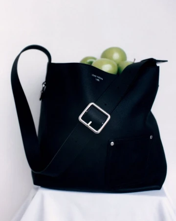 Black leather shoulder bag with a silver buckle and strap holding green apples against a white seamless background