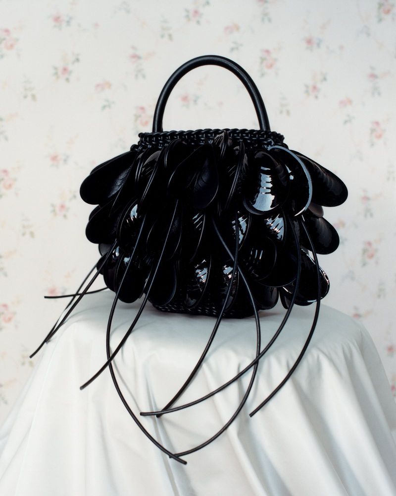 Black sculptural handbag with dangling petal-like attachments resting on a draped white fabric surface against floral wallpaper