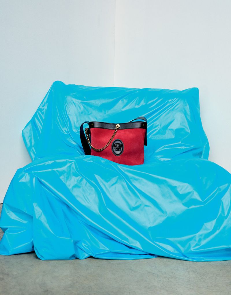 Red handbag with black leather trim resting on a bright blue crinkled plastic-covered furniture piece against a white wall