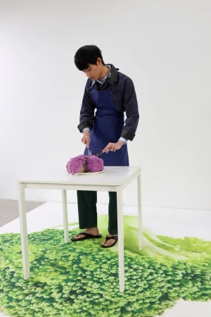 Man wearing a blue apron cutting purple cauliflower at a small white table over a green broccoli floor graphic