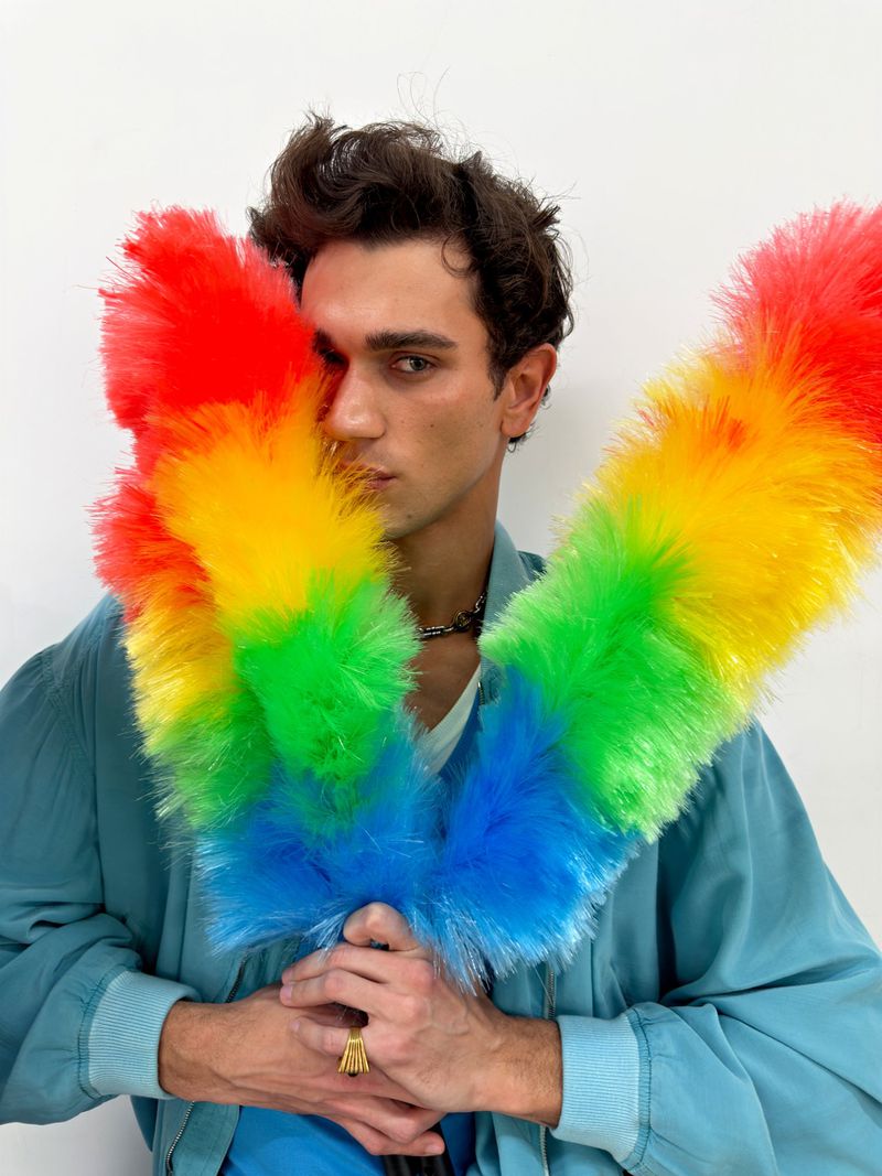 Man wearing a petrol blue jacket holding a colourful feathered duster in front of his face against a white background.