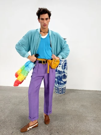 Model wearing a blue jacket, turquoise tank top, and purple trousers standing in a studio with a decorative vase.