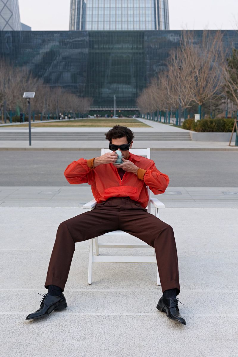 Model seated on a white chair outdoors wearing an orange shirt and brown trousers, drinking from a cup