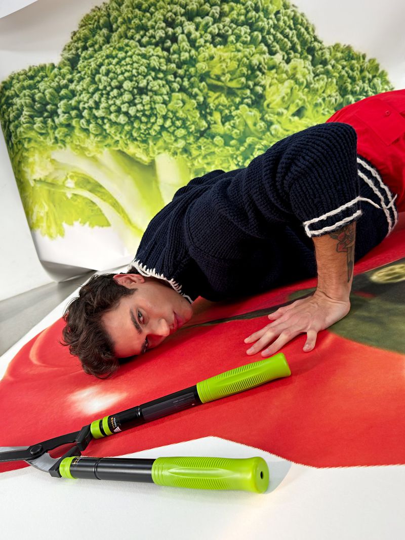 Adult male poses indoors on large printed images of produce; he wears a blue sweater, and oversized garden shears with green handles are in the foreground.