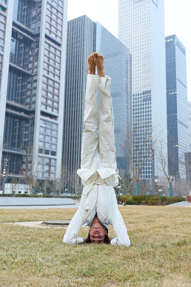 A man performing a headstand on a grassy lawn with tall modern skyscrapers rising in the background.