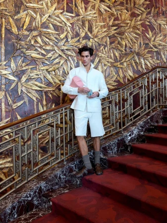 Male model in white jacket and shorts posing on a grand red carpeted staircase with an ornate golden textured wall backdrop.