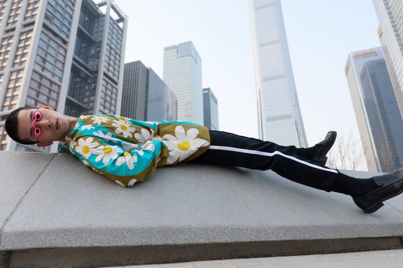 Male model wearing a floral print shirt and dark trousers reclining outdoors against an urban skyline backdrop.