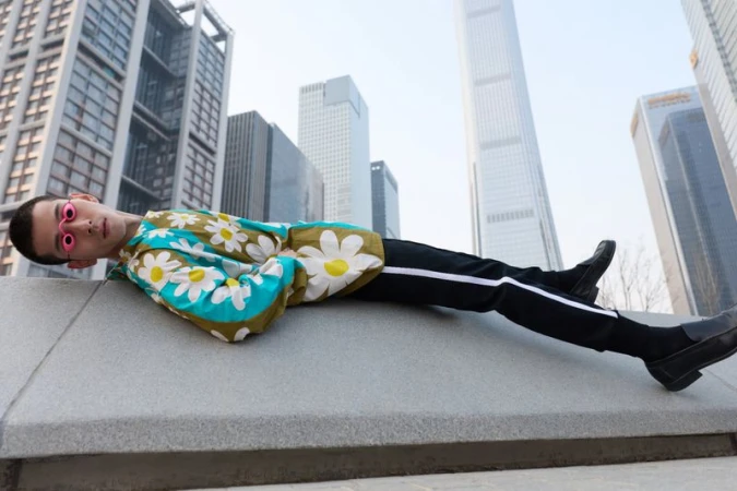 Male model wearing a floral print shirt and dark trousers reclining outdoors against an urban skyline backdrop.