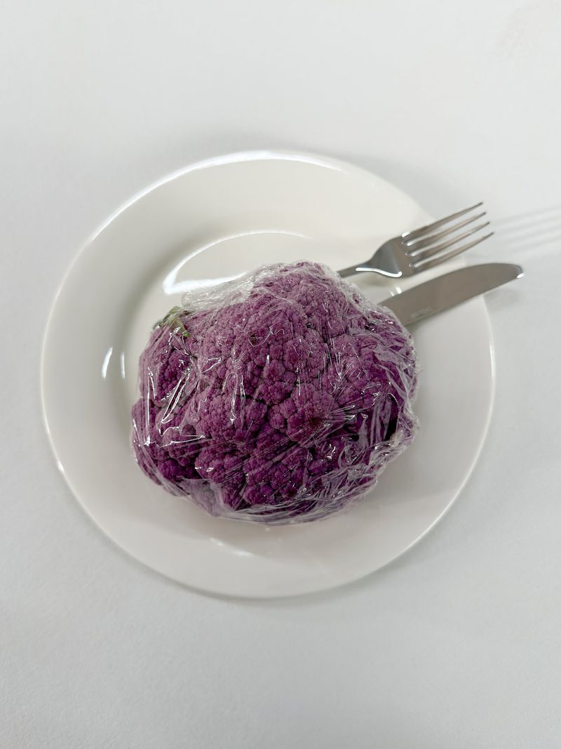 Top-down view of a wrapped purple cauliflower head on a white plate with a fork and knife on a white background.
