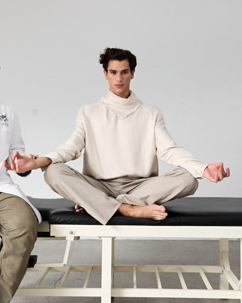 A man seated cross-legged in a meditative pose on a massage table against a neutral white background for a GQ China wellness editorial.