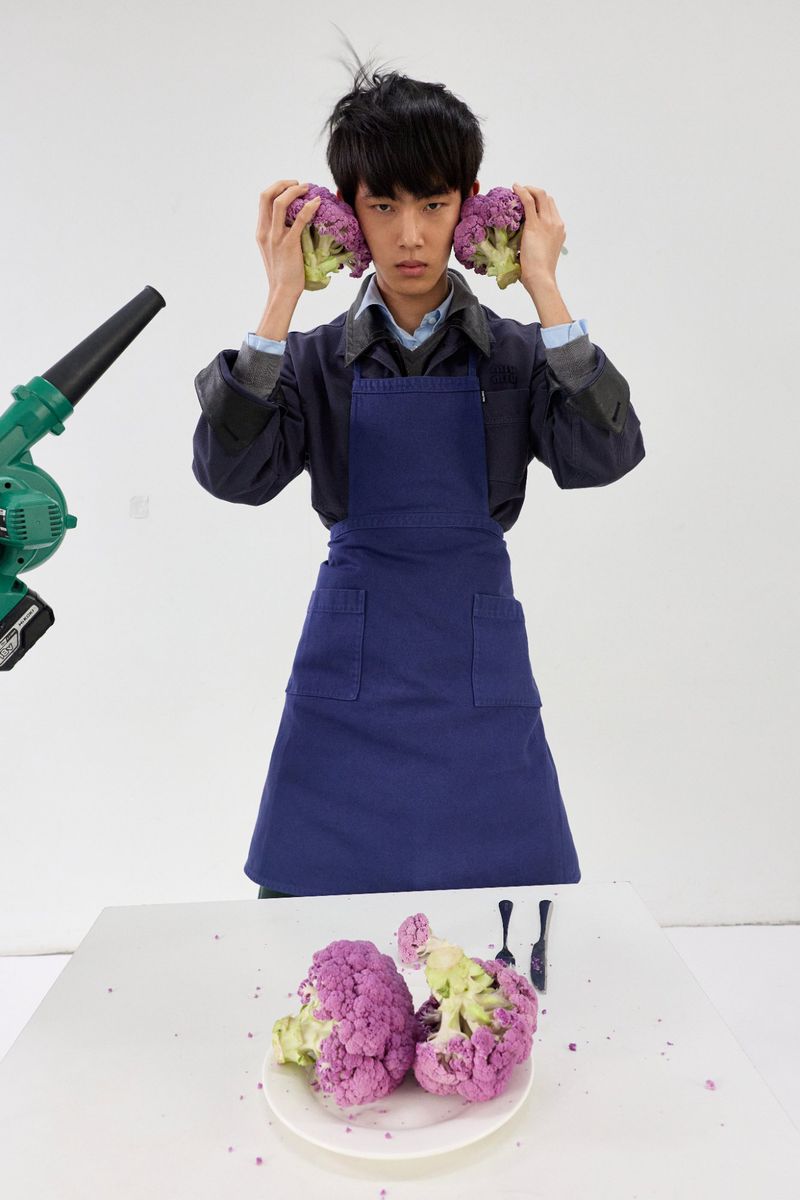 A man in a blue apron holds purple cauliflower florets to his ears against a white seamless background for GQ China.