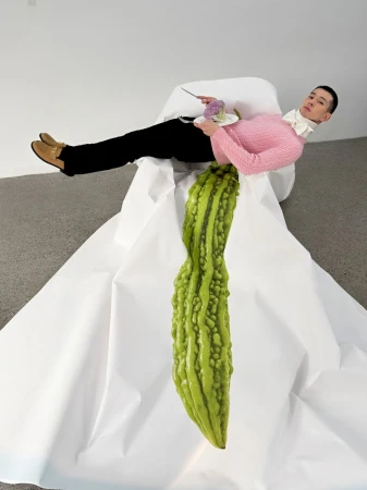 Model in pink sweater and black trousers lying on a large white paper installation with a giant green bitter melon