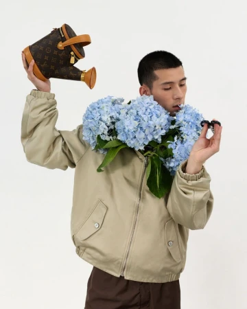 Man wearing a beige jacket holding a monogram watering can and blue hydrangeas against a white background.