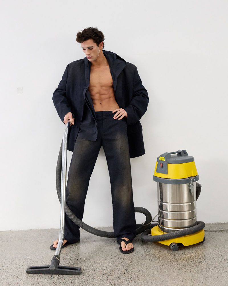 Shirtless male model wearing an oversized dark blazer and trousers standing next to a yellow vacuum cleaner in a studio