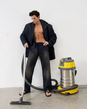Shirtless male model wearing an oversized dark blazer and trousers standing next to a yellow vacuum cleaner in a studio