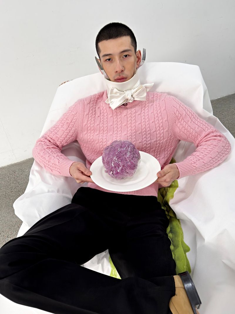 Man wearing a pink cable-knit sweater holding a plate with a purple object, seated against a white background for GQ China.