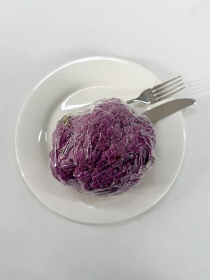 Top-down view of a wrapped purple cauliflower head on a white plate with a fork and knife on a white background.