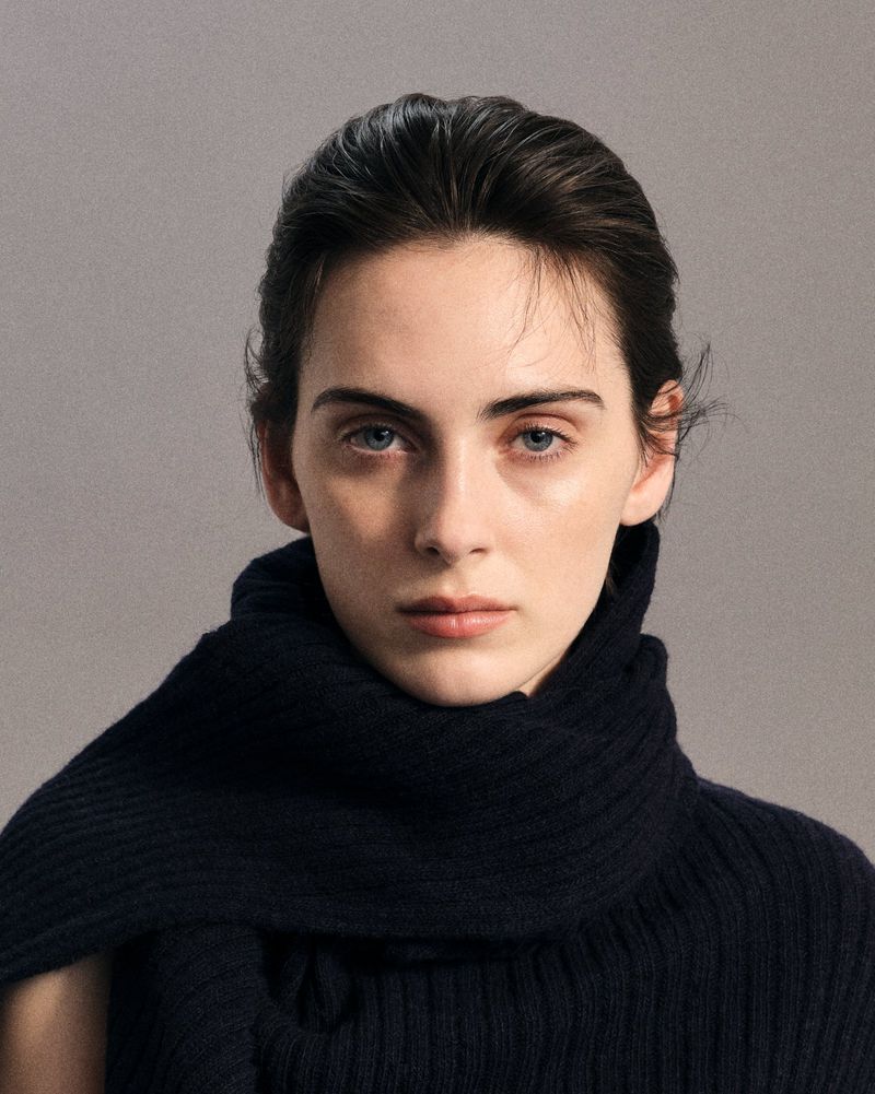 Portrait of a female model with slicked-back dark hair wearing a black knitted scarf against a neutral grey studio background
