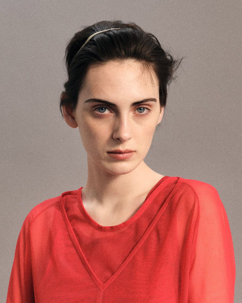 Portrait of a female model wearing a red top against a textured grey background for LUCA FABRY SS26 editorial.