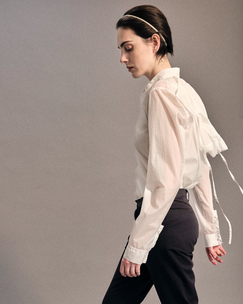 Model with dark hair wearing a white sheer button-down shirt and dark trousers against a plain grey studio background.