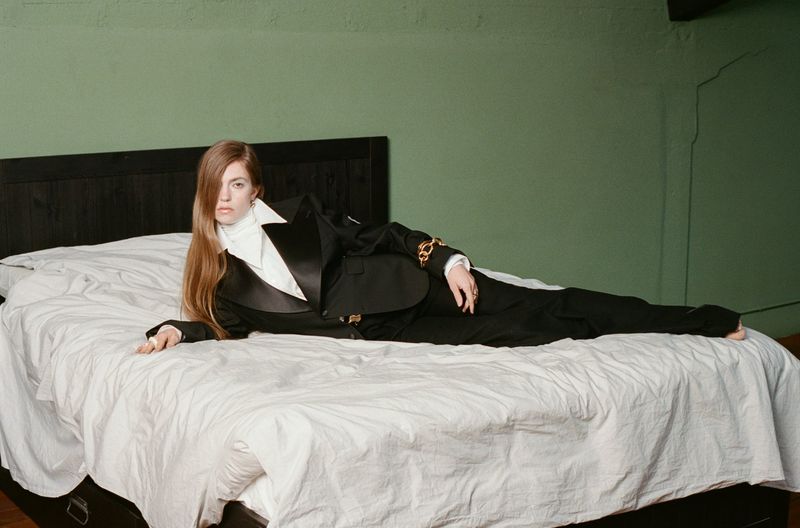 Woman in black tuxedo suit lying on a white bed against a green wall