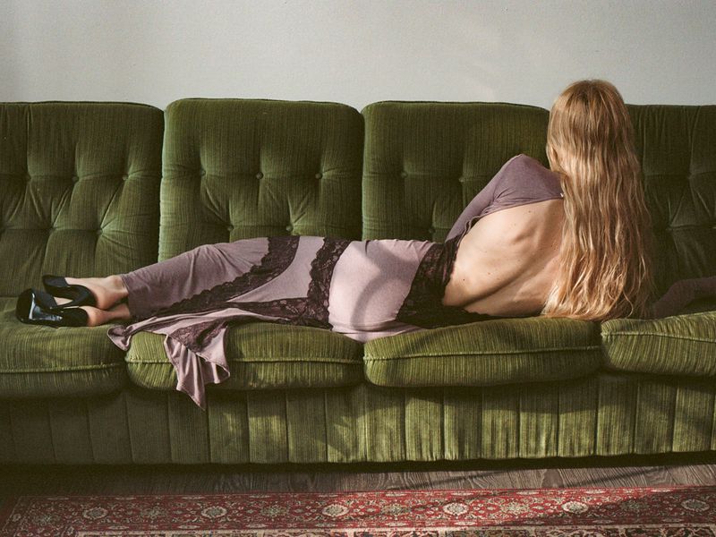 Woman with long blonde hair lying on a green tufted sofa wearing a mauve dress with lace detailing.