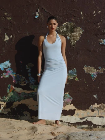 Model in a pale blue halterneck maxi dress standing barefoot against a weathered dark wall and sandy ground
