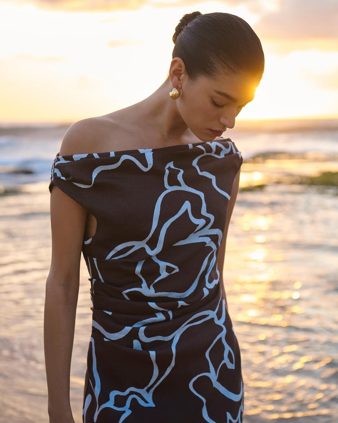 Model wearing dark patterned dress with gold earrings standing near ocean at sunset